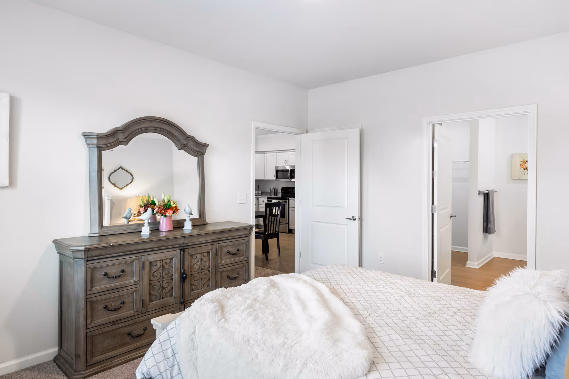 A bright bedroom with a large wooden dresser topped with a decorative mirror and flower arrangements. The bed is partially visible with a white and gray patterned comforter and a fluffy white pillow. Two open doors reveal a kitchen area with a dining table and a walk-in closet or bathroom area with towels hanging on the wall.