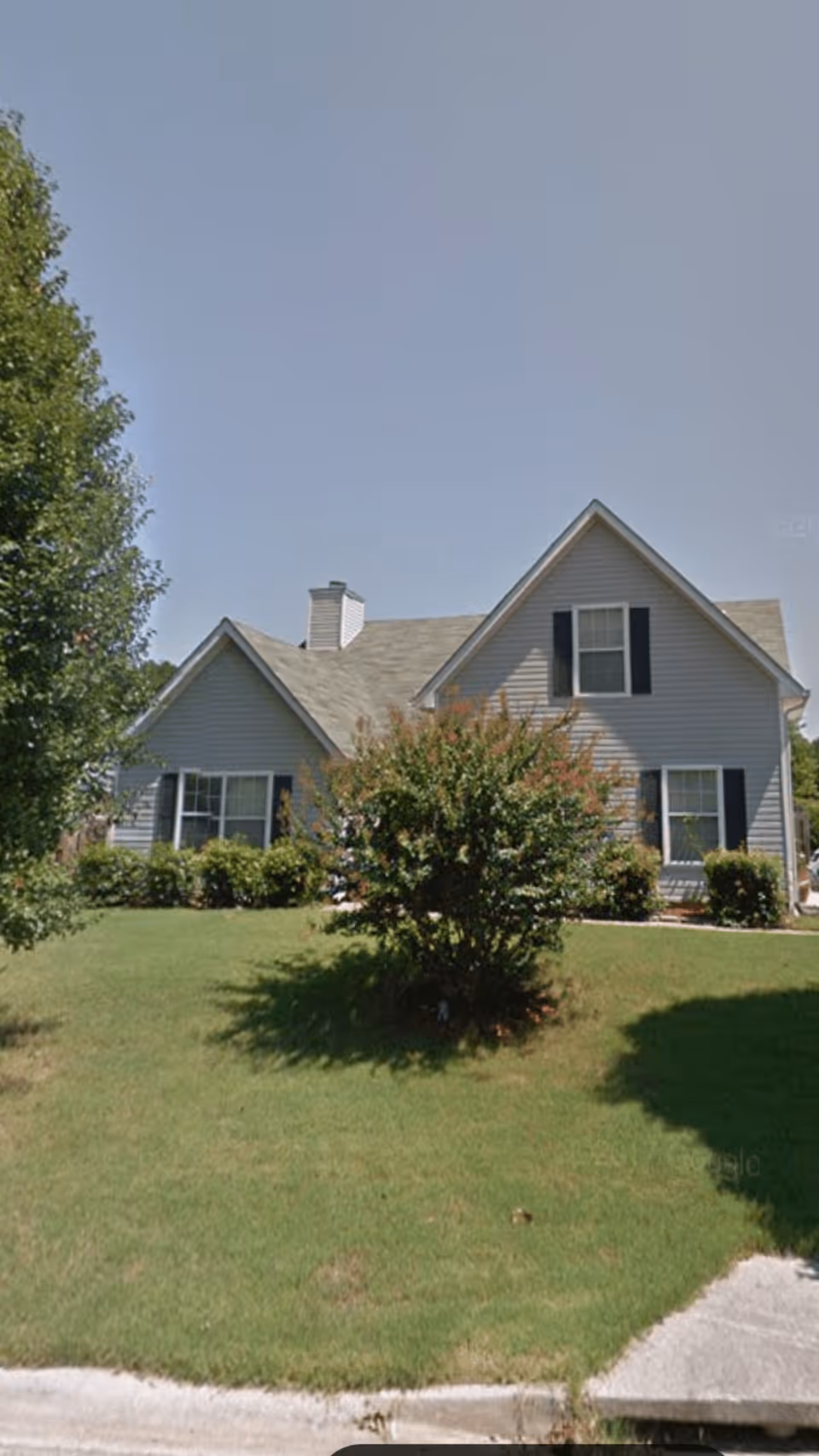 A single-story house with gray siding, black shutters, and a gabled roof. The house is surrounded by a well-maintained lawn with bushes and a tree in the front yard. The sky is clear and blue.