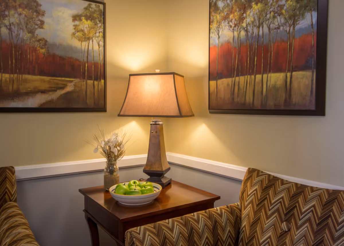 A cozy seating area with patterned armchairs, a wooden side table holding a lamp and a bowl of green apples, and framed landscape paintings on the wall.
