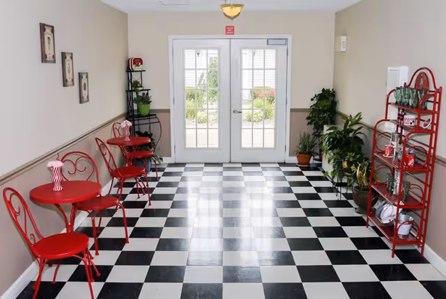 A bright interior space with black and white checkered flooring, featuring red metal tables and chairs on the left side, potted plants on the right side, and a red metal shelving unit holding glassware and dishes. Double glass doors at the far end lead outside to a garden area.