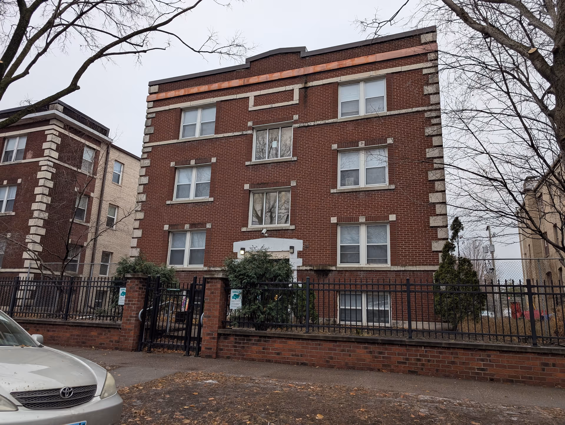 Three-story red brick apartment building with a black iron fence, leafless trees, and a parked car in front on a gray day.