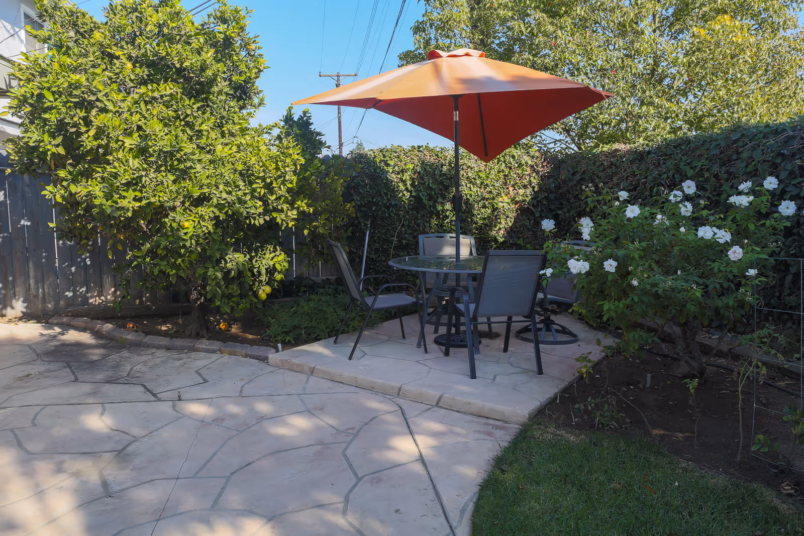Outdoor patio with a round glass table, four chairs and an orange umbrella surrounded by trees, flowering bushes and a stone walkway.