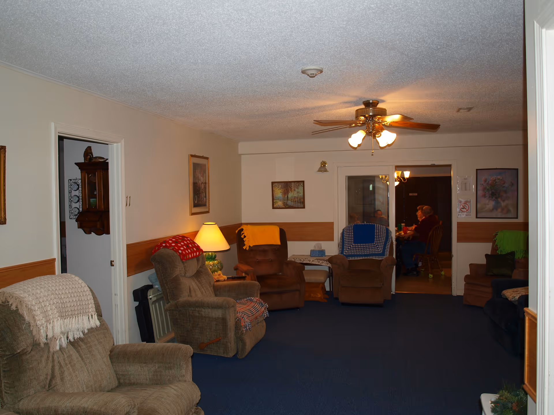 A cozy living room area in a senior living facility with several recliner chairs covered with colorful blankets. There is a ceiling fan with lights, a table lamp, framed artwork on the walls, and a doorway leading to a dining area where two people are seated at a table.