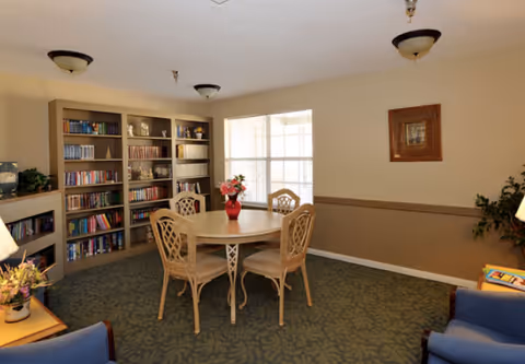 A cozy common area with a round wooden table and four chairs in the center. Behind the table is a large window letting in natural light. To the left, there is a built-in bookshelf filled with books and decorative items. The room has beige walls with a darker brown wainscoting and a green patterned carpet. Two blue armchairs are partially visible in the foreground, and a framed picture hangs on the wall.
