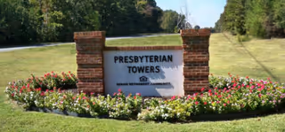 A brick-framed sign reading PRESBYTERIAN TOWERS sits on a manicured lawn with colorful flowers planted around it.