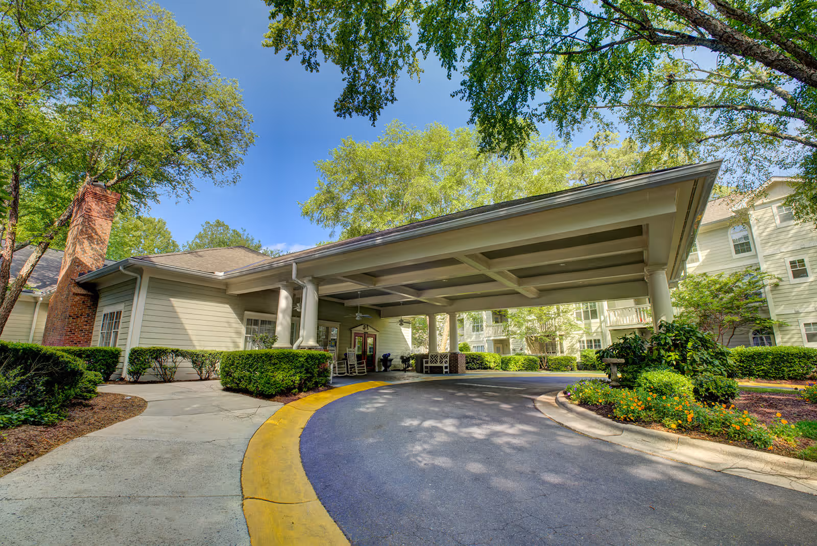 Covered driveway entrance of a senior living facility with a circular driveway, surrounded by green bushes and trees under a clear blue sky.