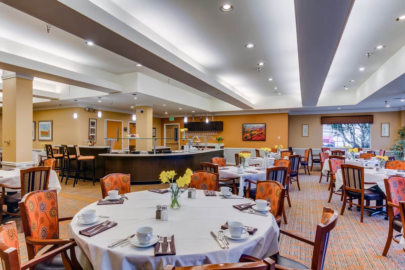A spacious dining room with round and rectangular tables covered with white tablecloths, set with cups, saucers, napkins, and silverware. The chairs have orange patterned upholstery. There are yellow flowers in vases on the tables. In the background, there is a counter area with bar stools and pendant lights hanging from the ceiling. The walls are painted in warm tones with framed artwork and a window letting in natural light.