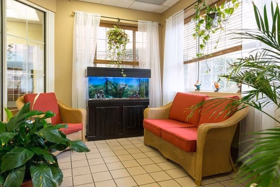 A cozy sitting area with two wicker chairs and a wicker loveseat, all with red cushions. There is a large fish tank on a black stand between the chairs, and several green plants around the room. The space is well-lit with natural light coming through large windows with white curtains and bamboo blinds.