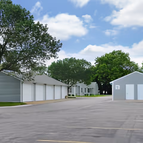 View of a paved driveway or parking area lined with single-story gray buildings with white doors, surrounded by green trees under a partly cloudy blue sky.