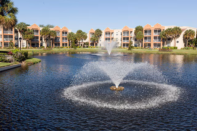 Fountain-filled pond in front of a multi-story building with palm trees under a clear sky.