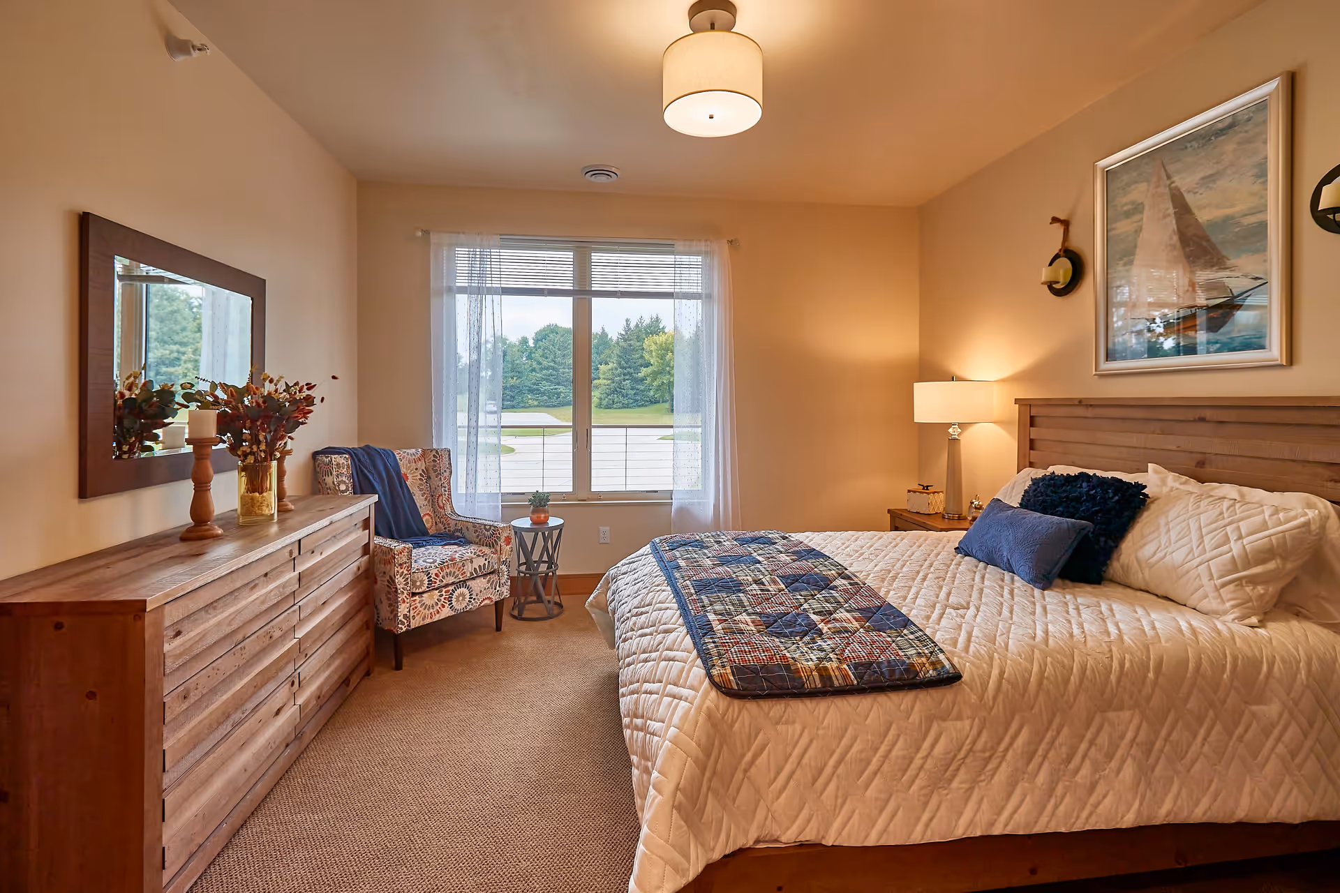 A cozy bedroom with a large bed covered in a white quilt and blue pillows. There is a wooden dresser with a mirror above it on the left side, and a patterned armchair next to a small round table by a window with sheer curtains. A framed picture of a sailboat hangs above the bed, and a lamp on a nightstand provides warm lighting.
