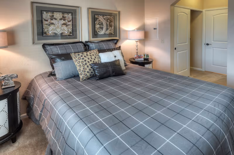 A neatly made bed with a gray checkered bedspread and multiple decorative pillows in a bedroom. Two framed artworks hang above the bed. There are two bedside tables with lamps on each side of the bed. The room has beige walls and carpeted floor, with a doorway leading to another room.