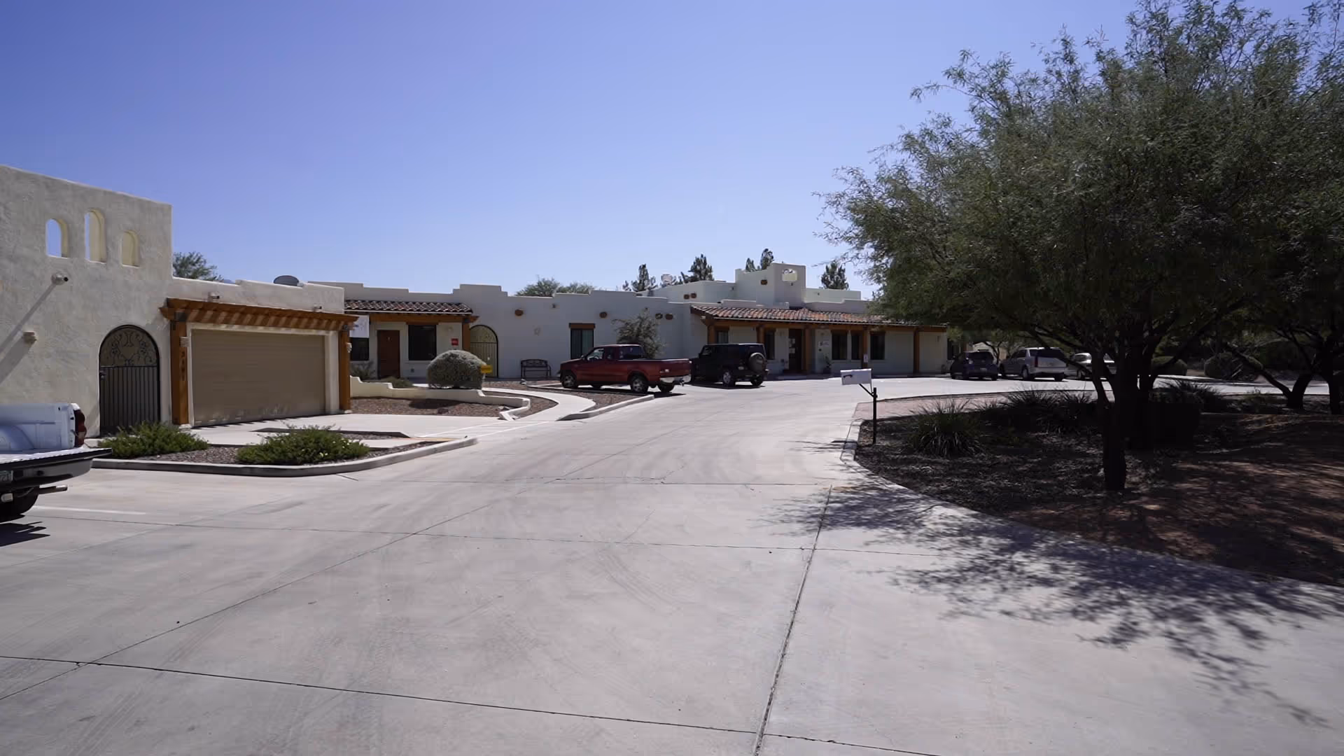 Exterior view of a single-story building with a southwestern architectural style, featuring white stucco walls, wooden beams, and a tiled roof. Several vehicles are parked in front of the building, and there are trees and landscaped areas along the driveway under a clear blue sky.