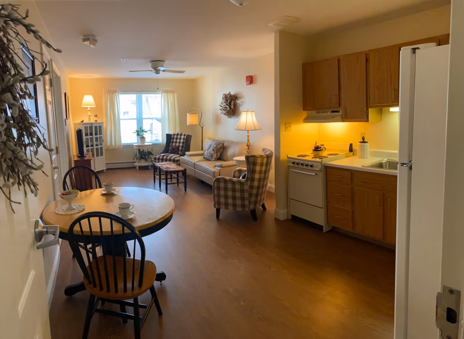 Interior view of a cozy retirement apartment featuring a small kitchen area with wooden cabinets, a stove, and a refrigerator on the right. In the center, there is a round wooden dining table with two chairs and two cups placed on it. The living area includes a beige sofa, two plaid armchairs, a small coffee table, a TV on a stand, and a window with light curtains letting in natural light. The room has warm lighting and wooden flooring.