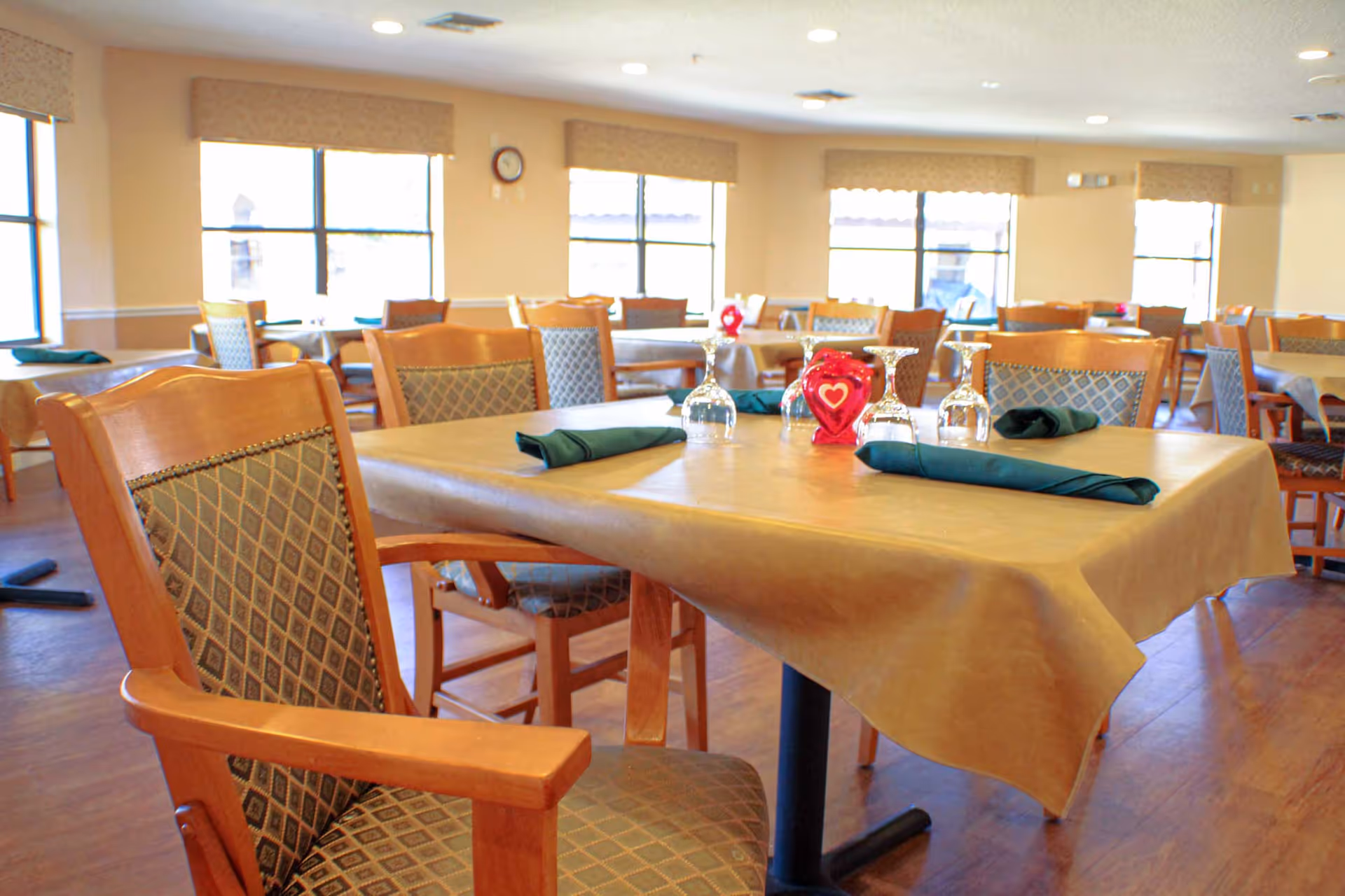 A bright dining room with multiple wooden tables and chairs. Each table is covered with a beige tablecloth and set with green cloth napkins and upside-down wine glasses. The room has large windows allowing natural light to fill the space.