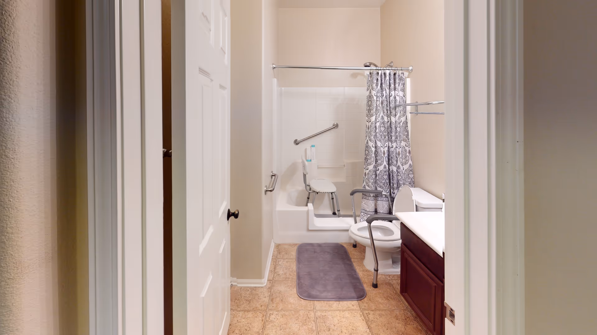 Accessible bathroom with a tub and shower chair, toilet with a support frame, vanity, and a patterned shower curtain.