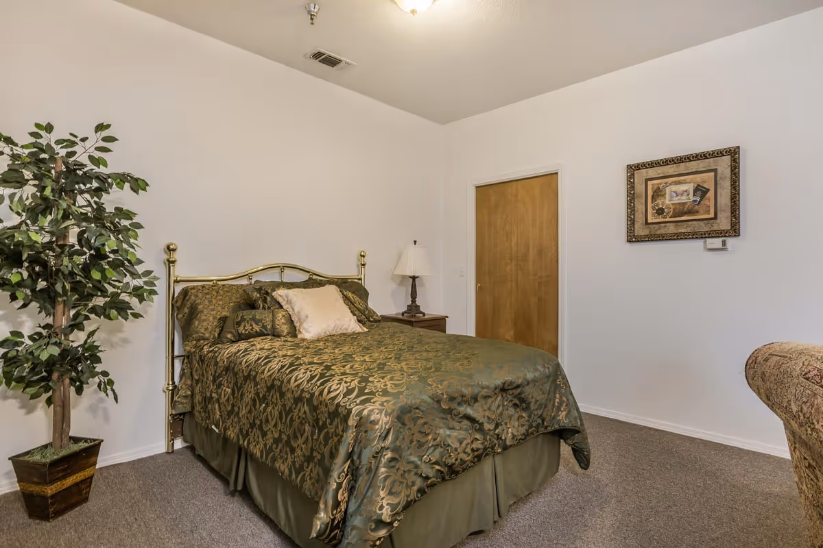 A bedroom with a bed covered in green and gold patterned bedding, a wooden nightstand with a lamp, a potted artificial plant, a closed wooden door, and a framed picture on the white wall.