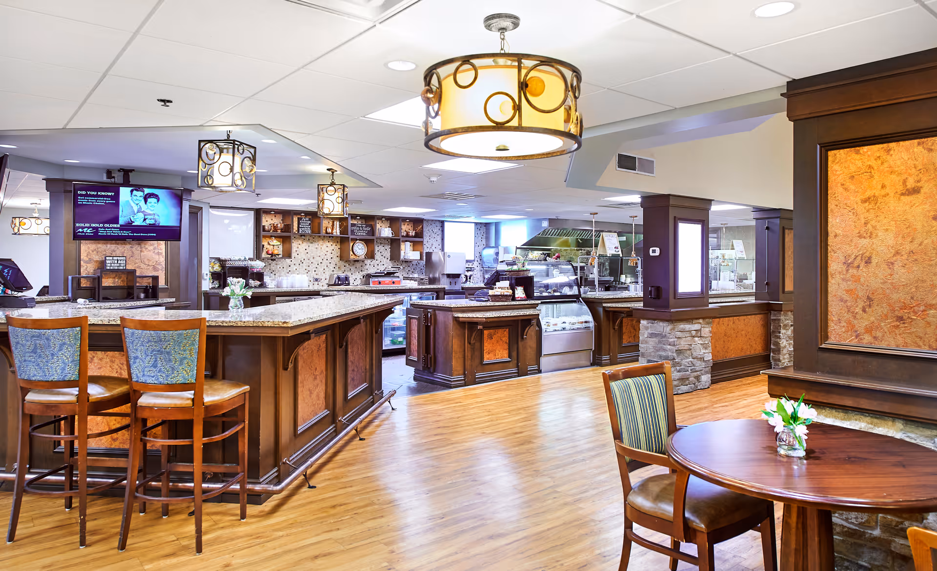 Interior view of a senior living facility dining area with a large wooden counter and bar stools, a round table with chairs, decorative hanging lights, and a kitchen area in the background with food display cases and appliances.