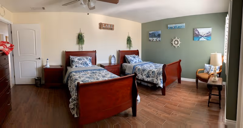 A bedroom with two wooden twin beds featuring blue and white floral bedding. Between the beds is a wooden nightstand with a decorative lantern. The walls are painted in light yellow and green, decorated with hanging plants, a wooden sign that says 'LAKE', and nautical-themed artwork including a ship's wheel. There is a wooden dresser on the left, a chair with a pillow, a small table with a lamp, and a ceiling fan above.