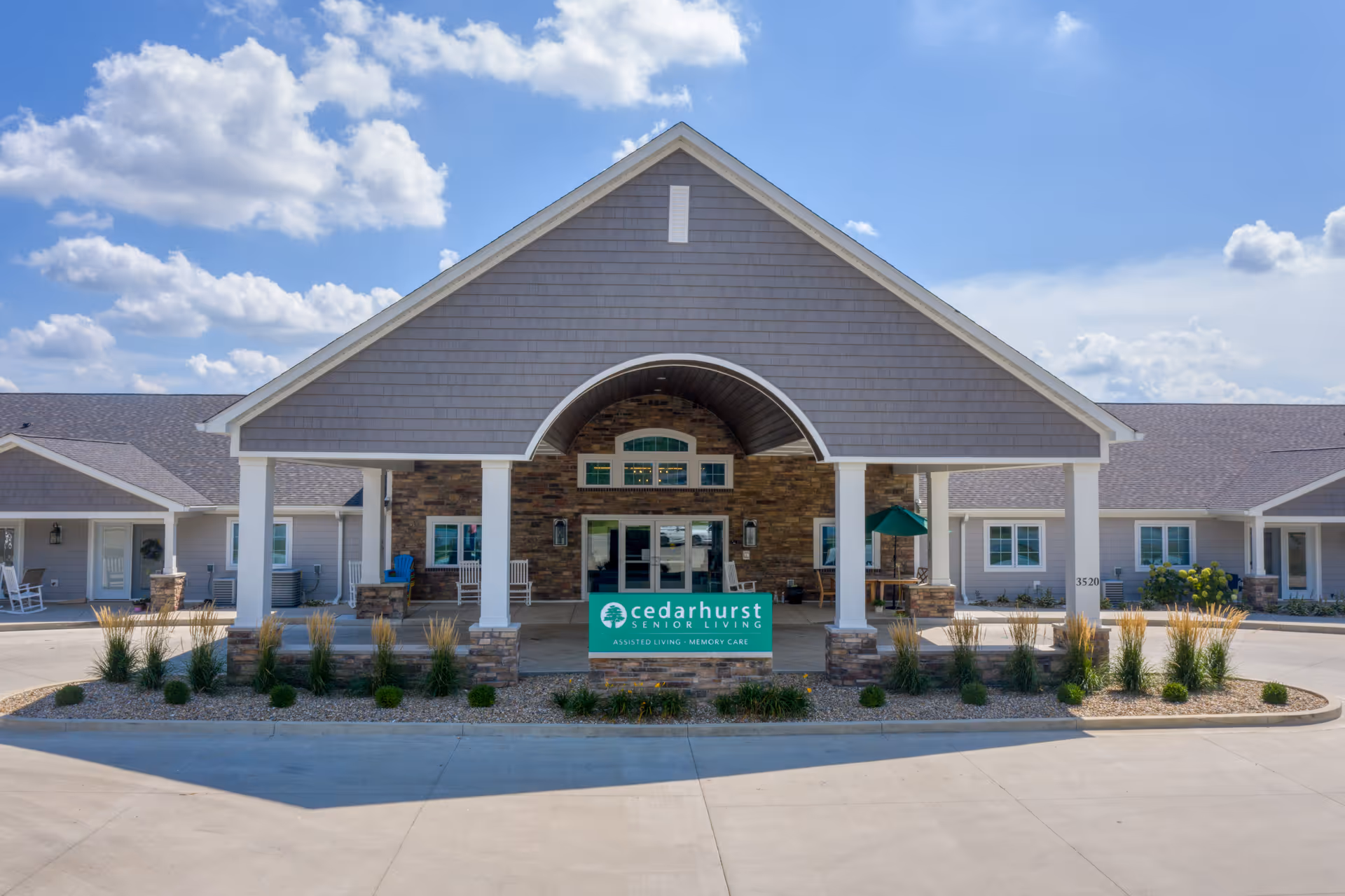Front exterior view of Cedarhurst Senior Living of Springfield, MO, showing a large covered entrance with stone and wood accents, landscaping with plants and shrubs, and a clear blue sky with some clouds.