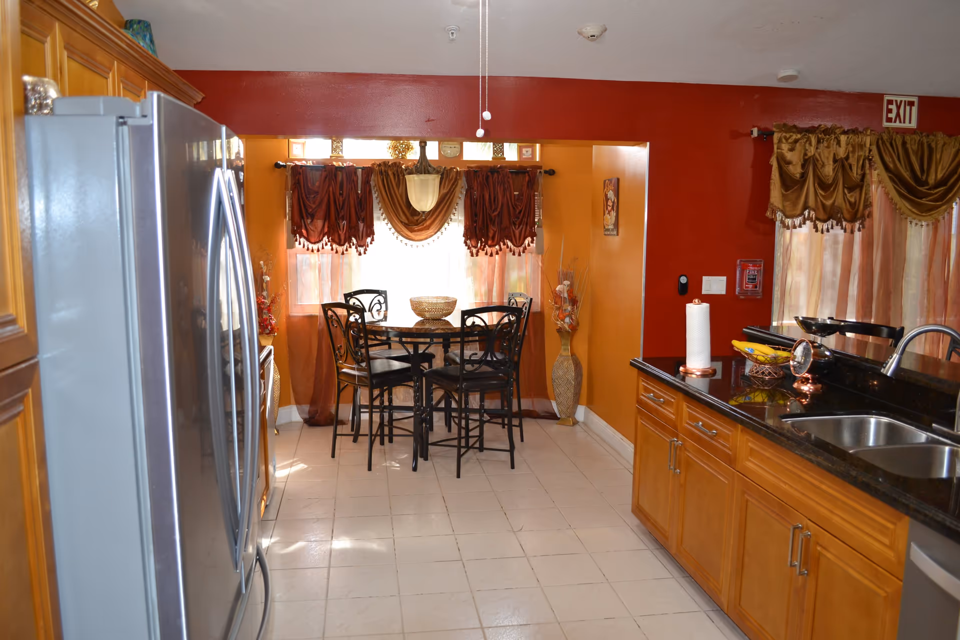 A kitchen and dining area with wooden cabinets, a stainless steel refrigerator, a black countertop with a sink, and a small dining table with four chairs near a window with decorative curtains. The walls are painted in warm tones of red and orange.