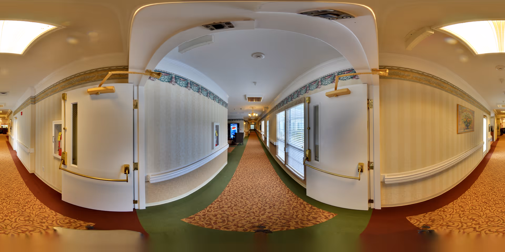 A long, carpeted hallway in a senior living facility with patterned wallpaper, white doors with brass handles, and large windows on one side letting in natural light. The hallway is decorated with framed artwork and has handrails along the walls.