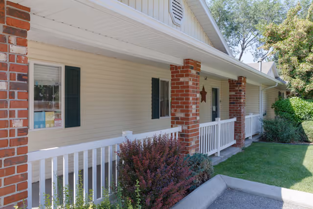 Covered front porch of a single-story building with brick columns, white railing, green shutters, and a small lawn.
