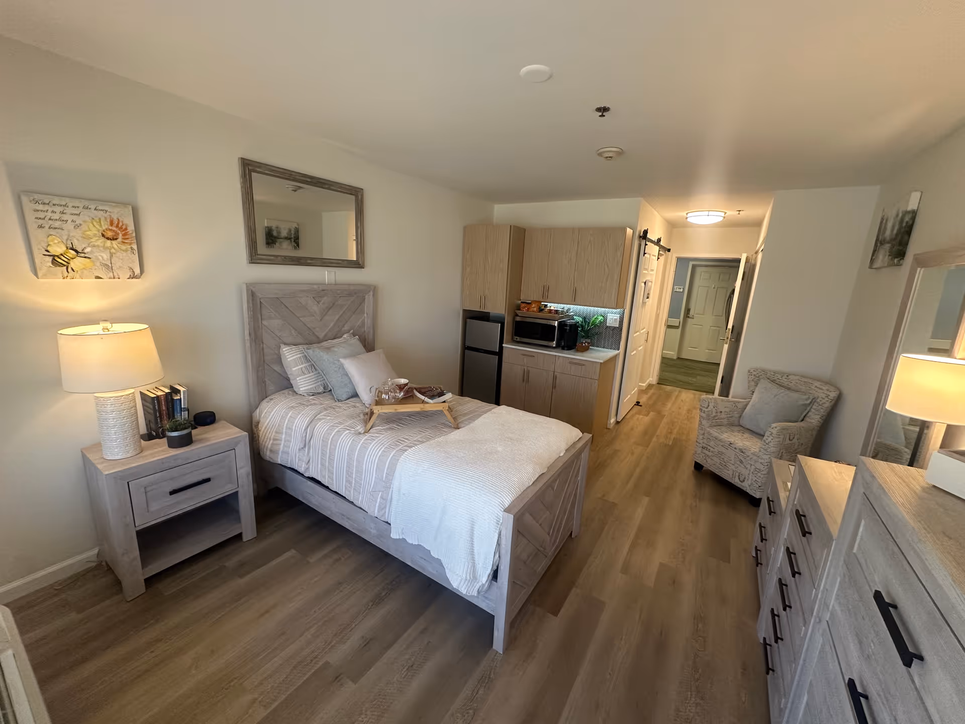 A cozy assisted living bedroom featuring a single bed with a light-colored wooden headboard and bedding, a bedside table with a lamp and books, a small kitchenette with a mini fridge, microwave, and cabinets, a comfortable armchair, and a dresser with a mirror. The room has wood flooring and soft lighting, creating a warm and inviting atmosphere.