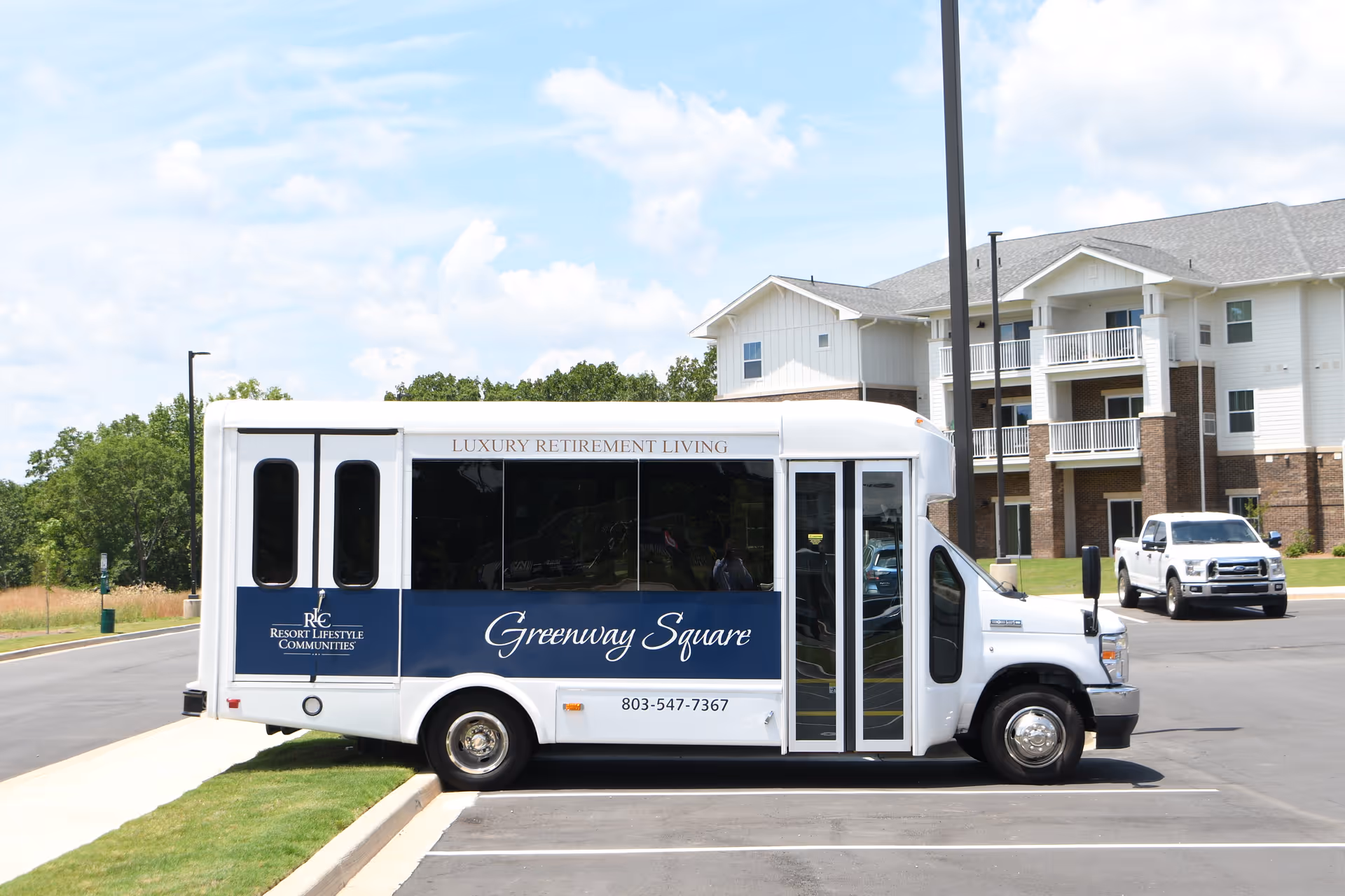 A white shuttle bus parked in a parking lot in front of a multi-story residential building. The bus has signage that reads 'Greenway Square' and 'Luxury Retirement Living' along with a phone number. The building has balconies and a brick and white exterior. A white pickup truck is also visible in the background.