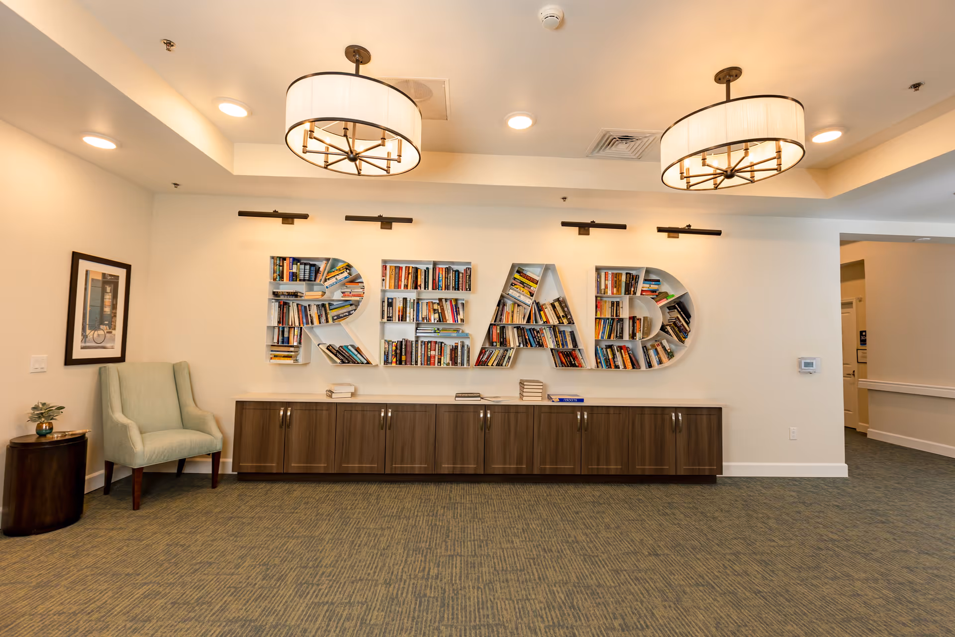Interior view of a reading area in a senior living facility with large wall-mounted bookshelves shaped as the word READ filled with books. Below the shelves is a long wooden cabinet. To the left is a light green armchair next to a small round table with a plant and a framed picture on the wall. The ceiling has two modern circular light fixtures.