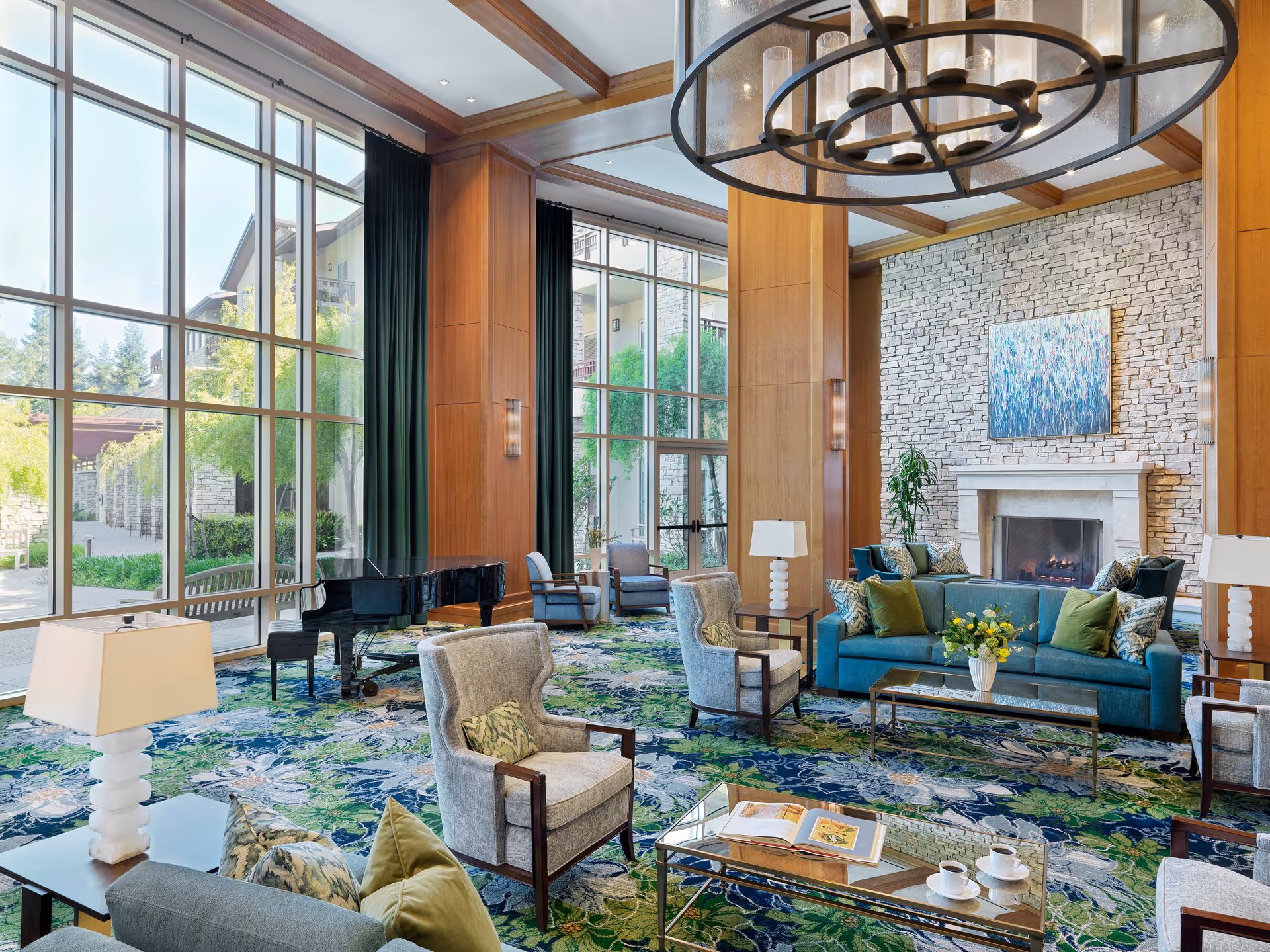 A spacious and elegant living room with large floor-to-ceiling windows letting in natural light. The room features a stone fireplace with a colorful painting above it, a teal sofa with green and patterned pillows, several gray armchairs, a glass coffee table with an open book and two cups, and a black grand piano near the windows. The carpet has a floral pattern in shades of blue and green, and there are modern lamps on side tables. Outside the windows, greenery and parts of the building exterior are visible.