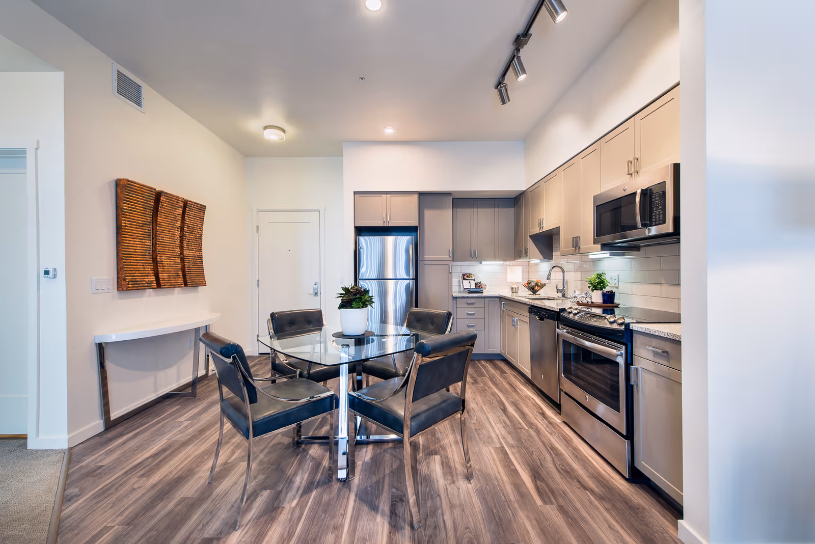 Modern kitchen and dining area with stainless steel appliances including a refrigerator, stove, microwave, and dishwasher. The kitchen has light gray cabinets, white subway tile backsplash, and granite countertops. A round glass dining table with four black chairs is placed on wood flooring. A small potted plant is on the dining table, and a decorative wooden wall art hangs on the adjacent wall.