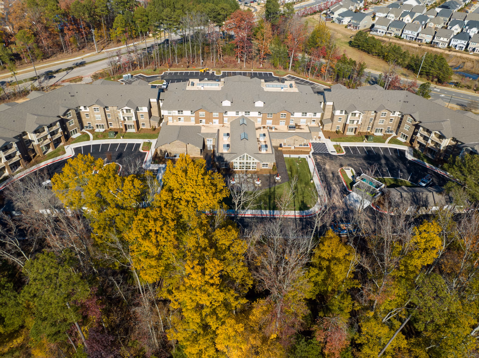 Aerial view of Brier Pointe Retirement Community showing a large multi-wing building surrounded by parking lots and autumn-colored trees. The building has a gray roof and beige exterior walls, with a central courtyard area featuring outdoor seating and greenery.
