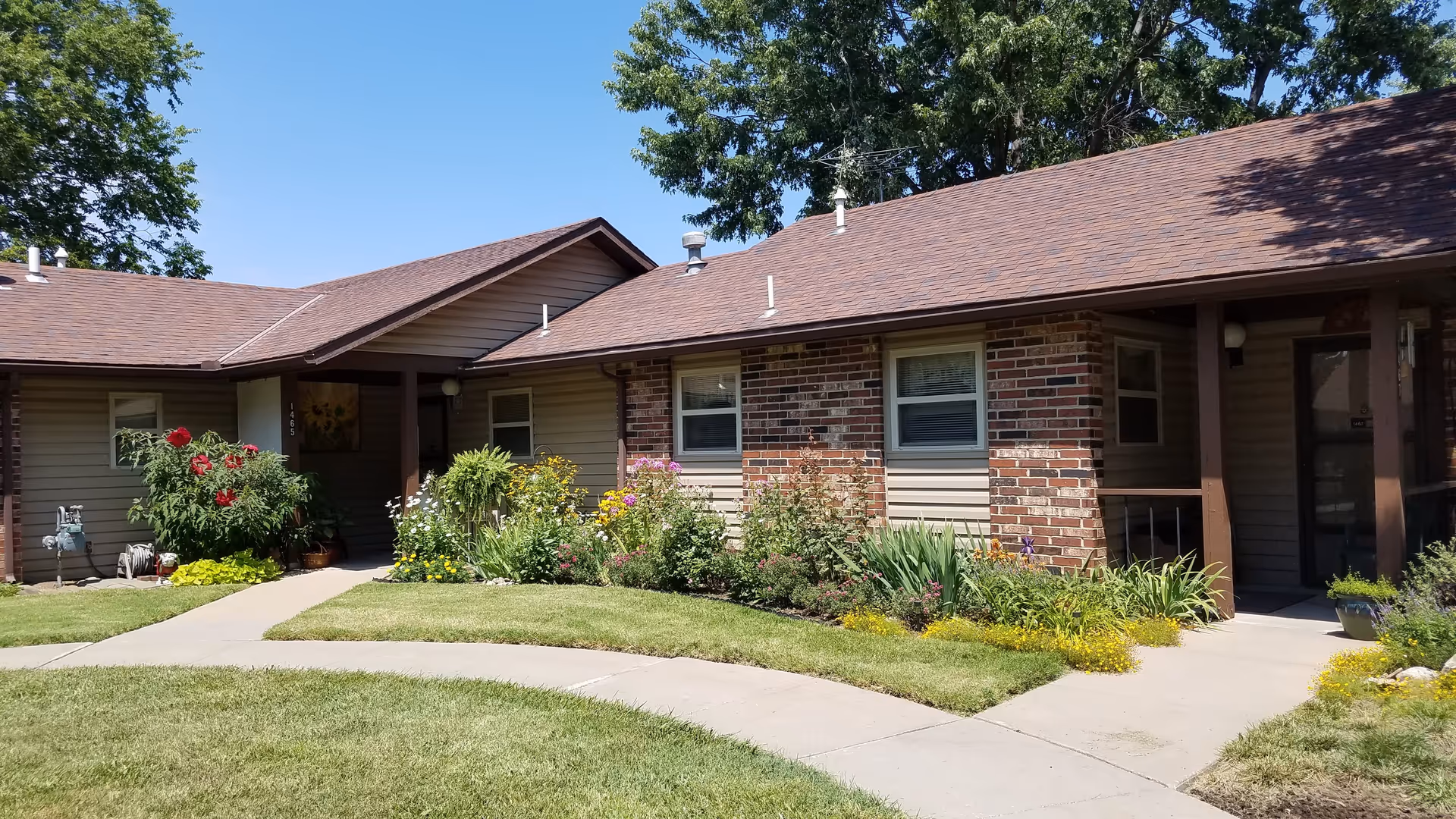 Single-story brick-and-siding senior living building with flower beds and a curved sidewalk in front under a clear blue sky.