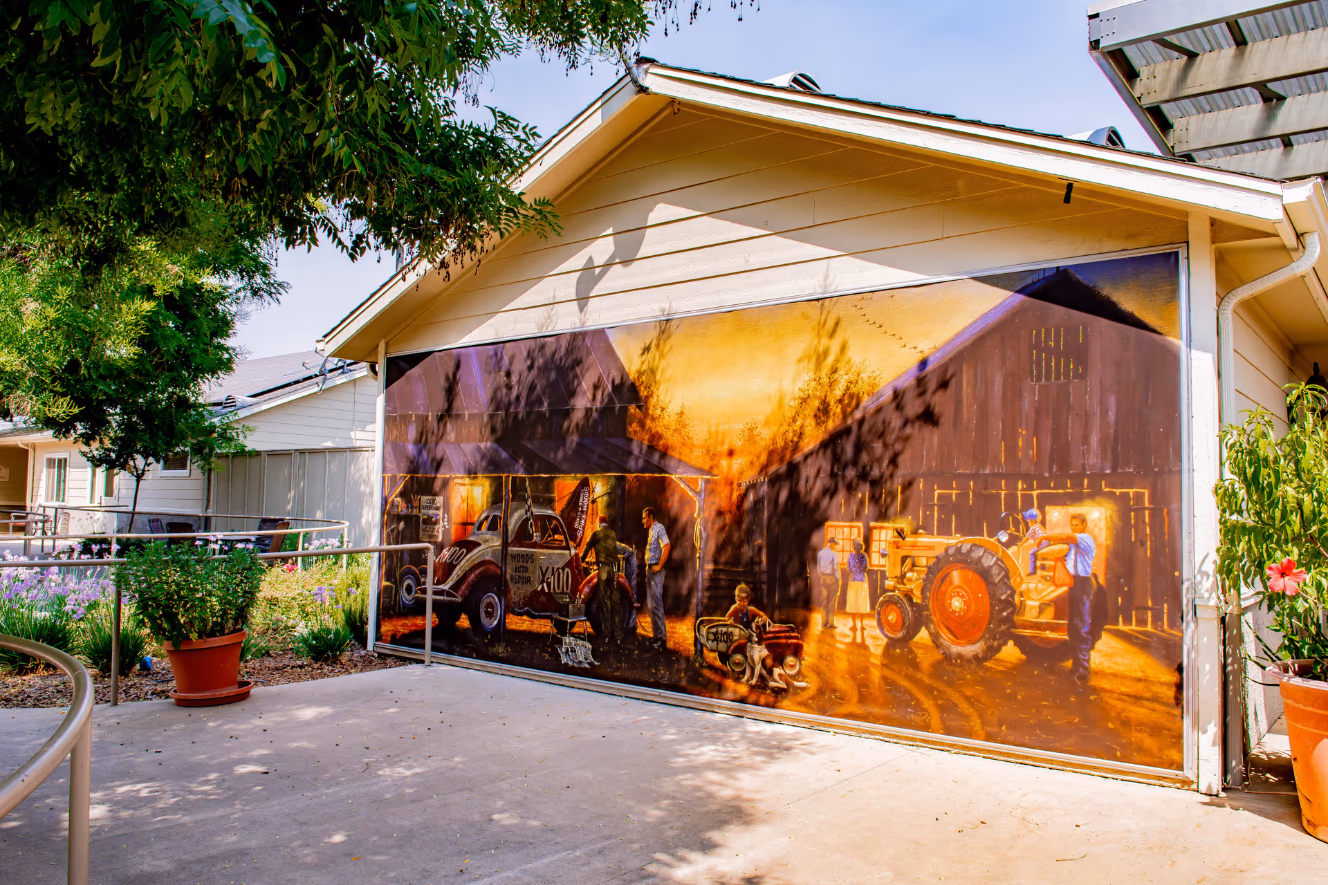 Exterior view of a building with a large mural painted on the side wall depicting a vintage farm scene with people, a tractor, and an old car in warm sunset colors. There are plants and trees around the building and a concrete pathway with a metal railing in front.