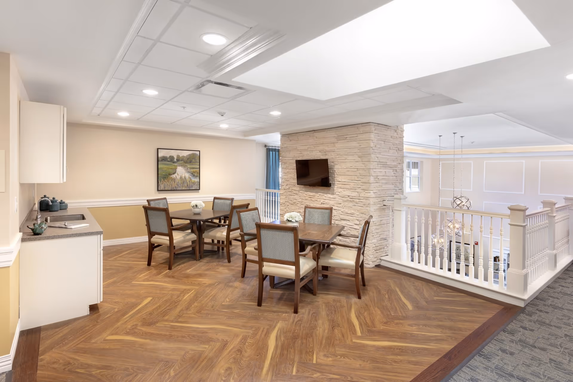 A bright and spacious common area in an assisted living facility featuring two wooden tables each surrounded by four cushioned chairs. The room has a wood-patterned floor, a stone accent wall with a mounted flat-screen TV, a small kitchenette with a countertop and cabinets, and a large skylight overhead. The area overlooks a lower level with white railing and modern pendant lights.