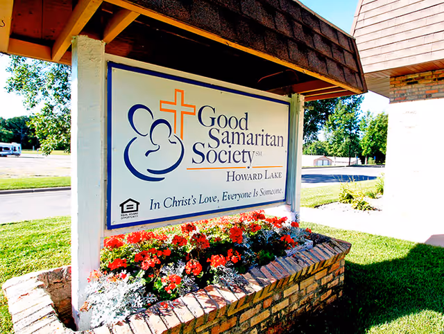 Outdoor sign for Good Samaritan Society Howard Lake with a cross and logo, surrounded by a flower bed with red and white flowers, next to a brick building and green lawn.