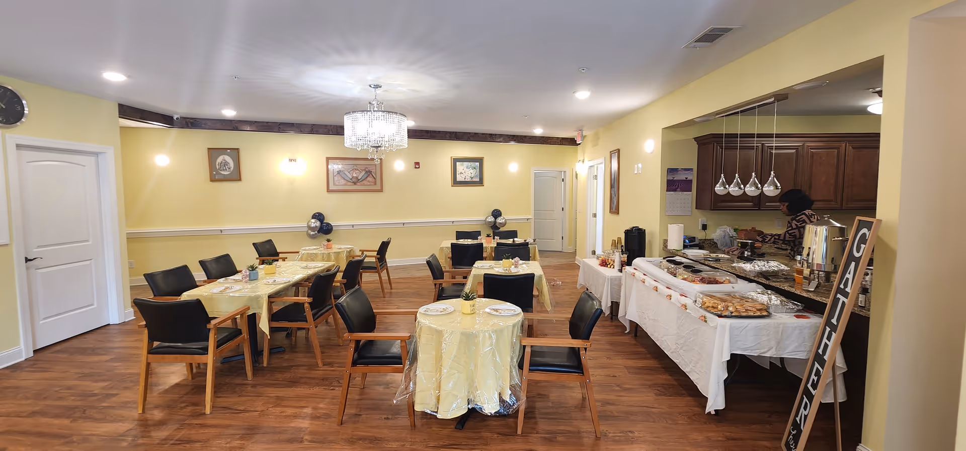 A dining room in a senior care facility with several tables covered in yellow tablecloths and set with plates and utensils. Black chairs surround the tables. On the right side, there is a buffet table with various food items and a person serving themselves. The room has yellow walls, wooden flooring, framed pictures on the walls, and a chandelier hanging from the ceiling. A sign with the word 'GATHER' is placed near the buffet table.