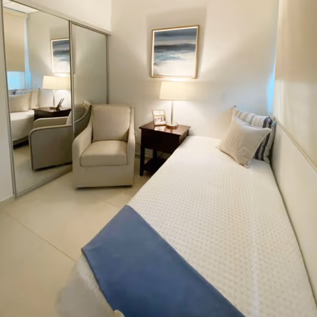 A small, cozy bedroom with a single bed covered in a white textured bedspread and a blue throw blanket at the foot. There is a beige armchair next to a dark wooden nightstand with a lamp and a framed photo. A large mirror closet door reflects part of the room. A framed abstract painting hangs on the wall above the nightstand.