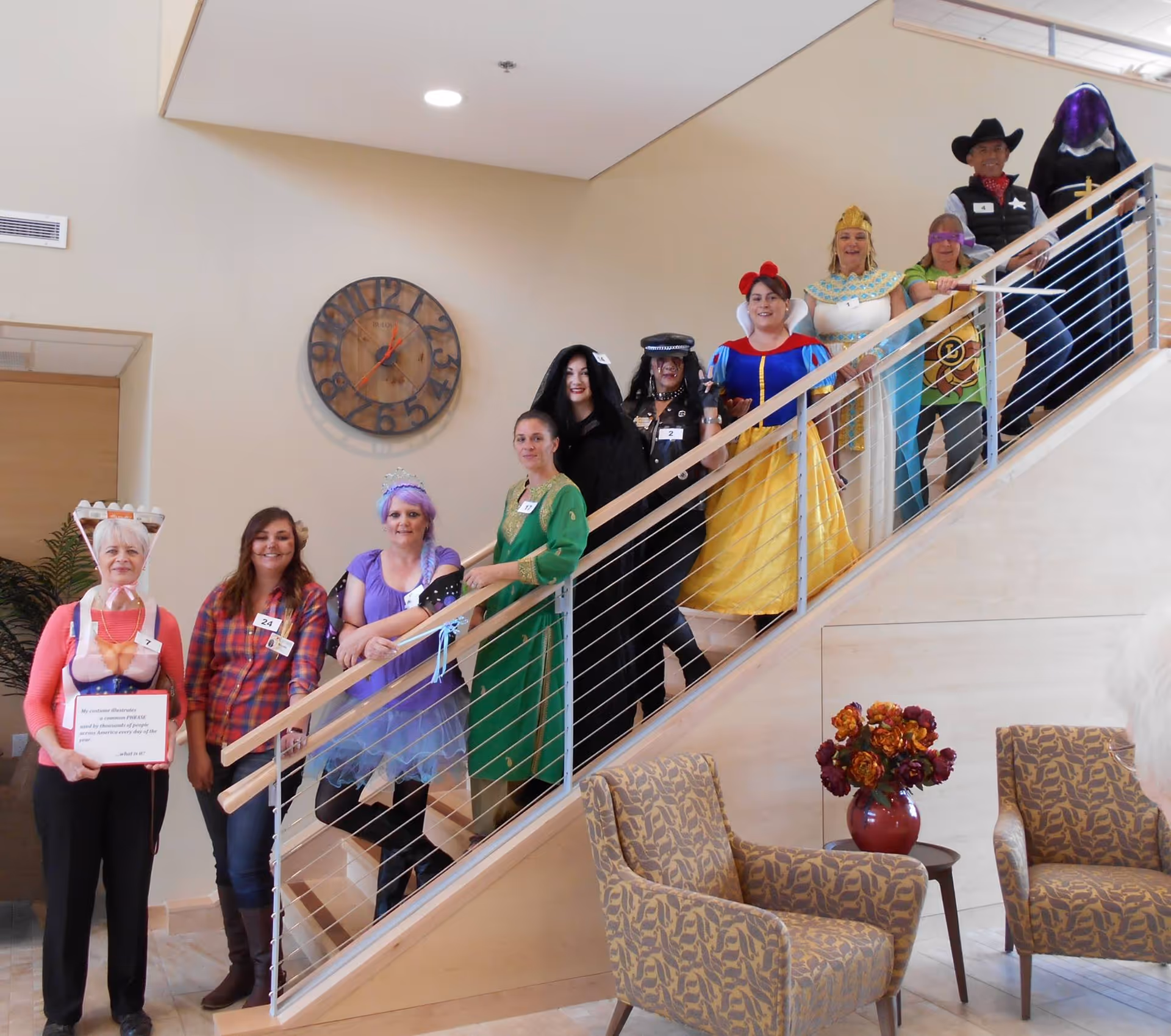 A group of people dressed in various costumes standing on a staircase inside a building. The costumes include a queen, a police officer, Snow White, an Egyptian queen, a cowboy, and others. The interior features beige walls, a large round clock, patterned armchairs, and a vase with flowers on a small table.