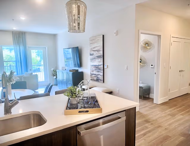 Open-plan apartment with a kitchen island and sink in the foreground and a bright living room with TV and balcony doors in the background.