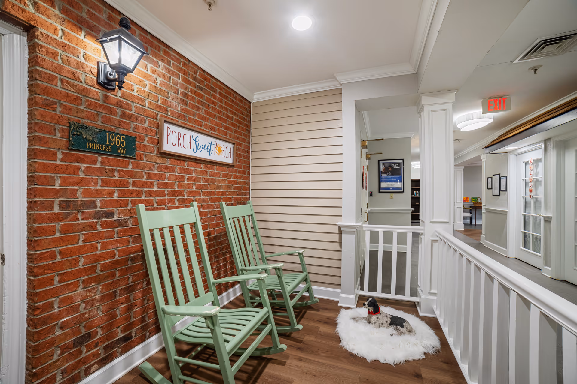Indoor porch area with two green rocking chairs against a red brick wall. A sign on the wall reads 'PORCH Sweet PORCH'. A small dog with a red collar lies on a white fluffy rug on the wooden floor. The area is well-lit with a wall-mounted lantern-style light and ceiling lights. There is a white railing and a hallway with an exit sign in the background.