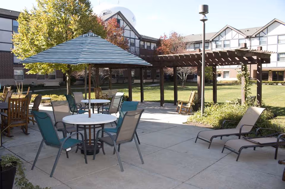 Outdoor patio area with several tables and chairs, including a round table with a striped umbrella, lounge chairs, and wooden rocking chairs. The patio is surrounded by a grassy lawn, trees with autumn foliage, and a wooden pergola. In the background, there is a multi-story building with large windows.