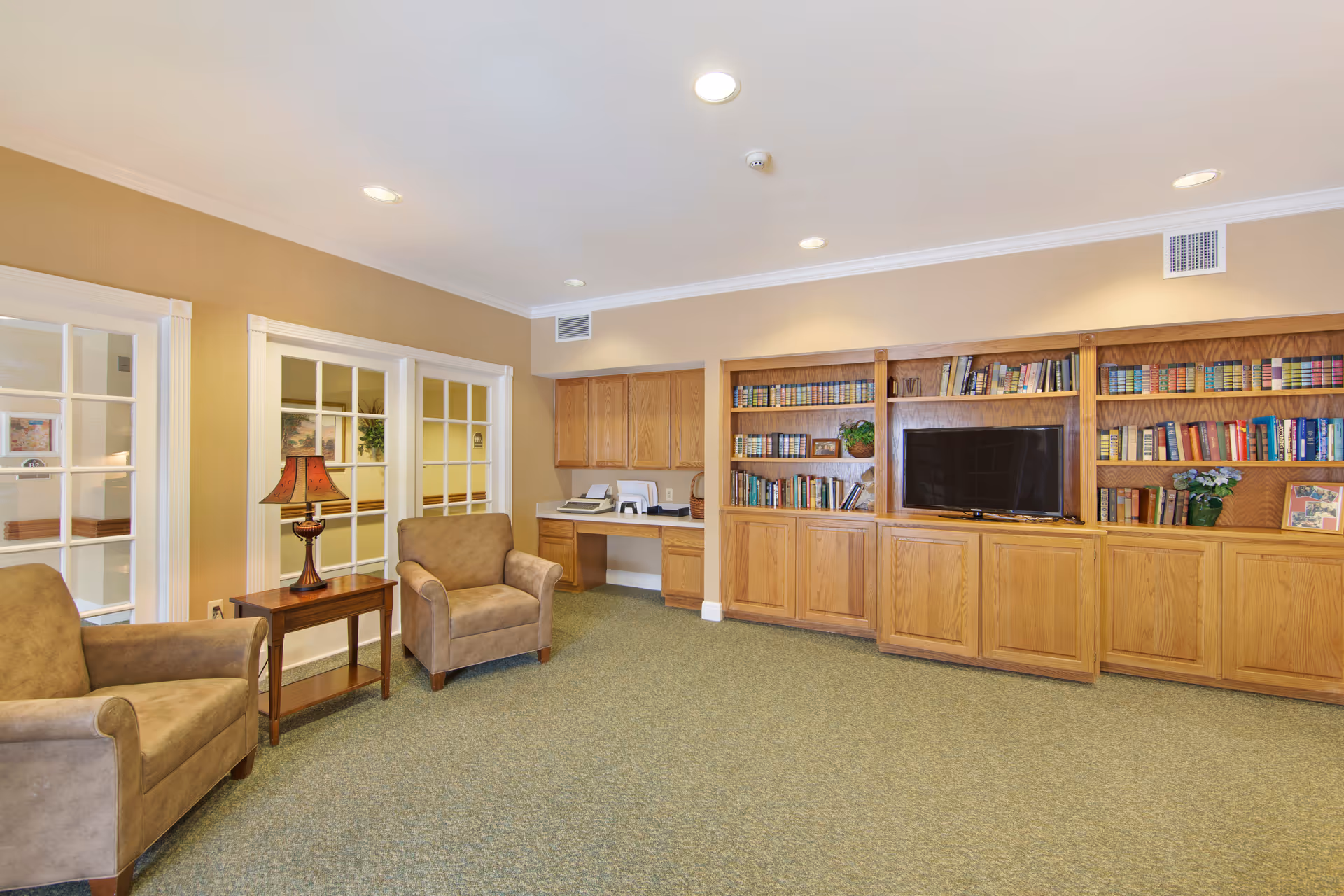 Well-lit lounge with upholstered chairs, wooden bookshelves and a TV in a senior living facility.