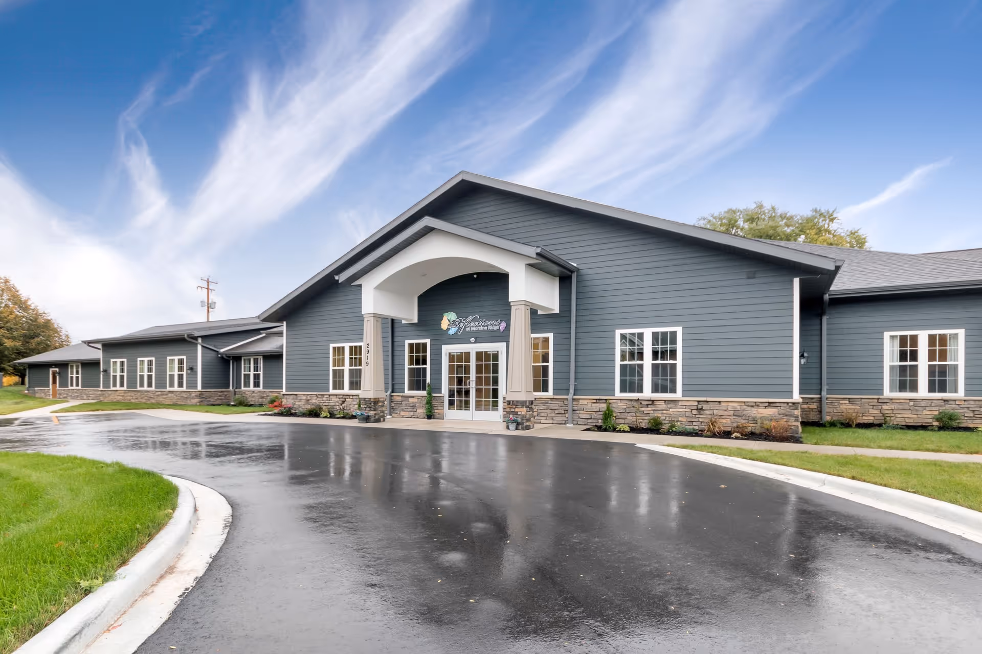 Exterior front view of Reflections at Moraine Ridge Memory Care facility, showing a single-story building with gray siding, stone accents, multiple windows, and a covered entrance with double glass doors. The driveway is wet, reflecting the building and the partly cloudy sky above.