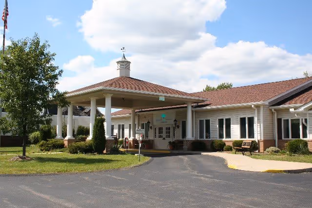 Exterior view of a single-story building with a covered entrance supported by white columns, surrounded by green grass and a tree under a partly cloudy sky.