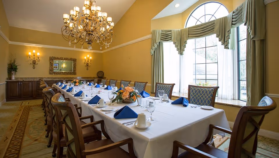 A formal dining room with a long rectangular table covered with a white tablecloth, set with blue folded napkins, glassware, and white cups. The room features large arched windows with green drapes, a chandelier hanging from a high ceiling, wall sconces, a mirror, and a sideboard with decorative items.