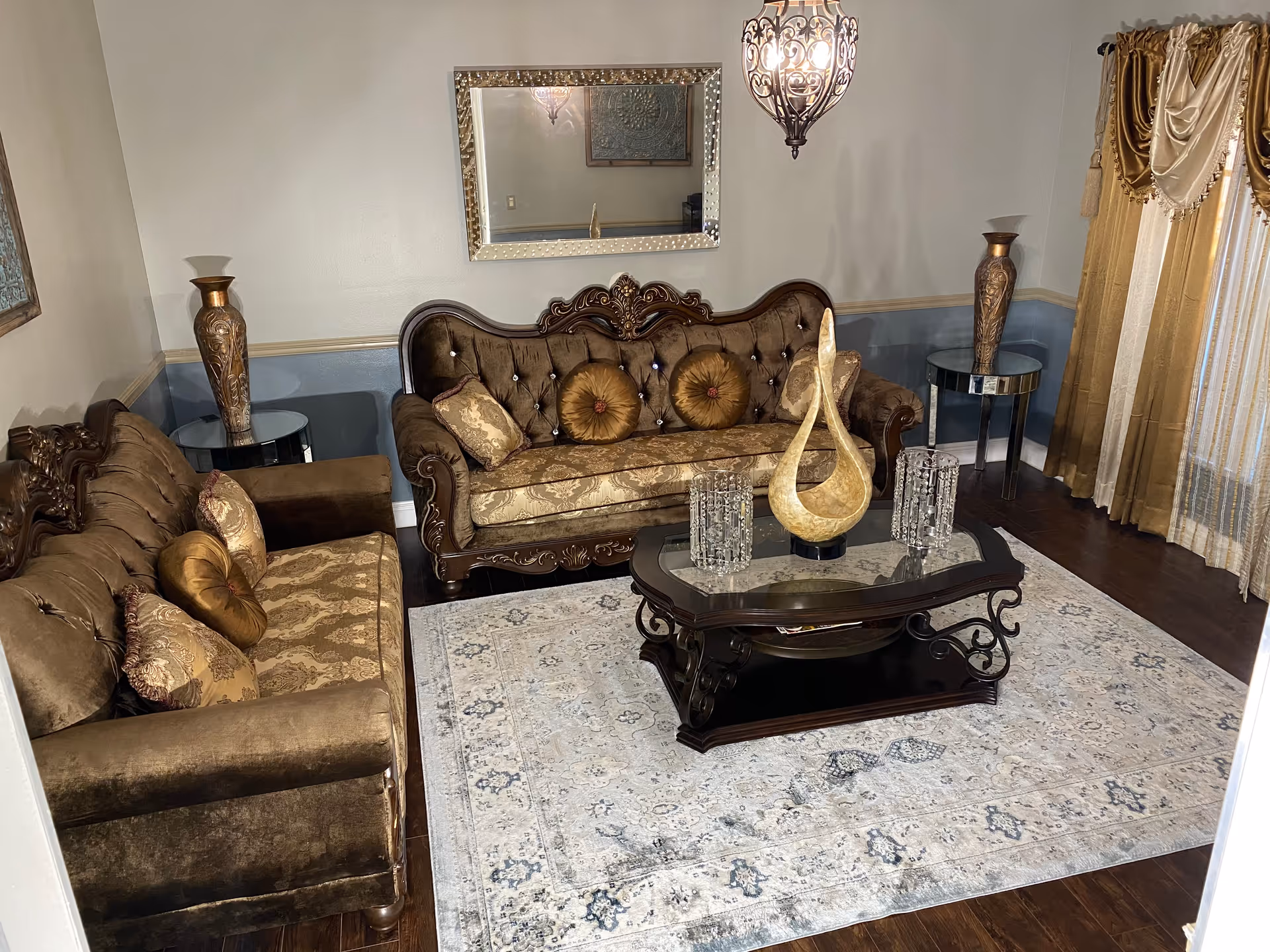 A living room with two ornate brown sofas adorned with patterned and round cushions, a dark wooden coffee table with decorative items on top, a large light-colored area rug with blue patterns, two side tables each holding a tall decorative vase, a large rectangular mirror on the wall, and gold and cream curtains covering a window.