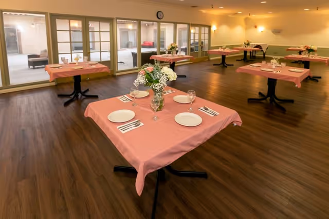 A dining room with several square tables covered with pink tablecloths, each set with white plates, silverware, and glasses. Each table has a vase with flowers as a centerpiece. The room has wooden flooring and large windows along one wall, with a clock above the windows.
