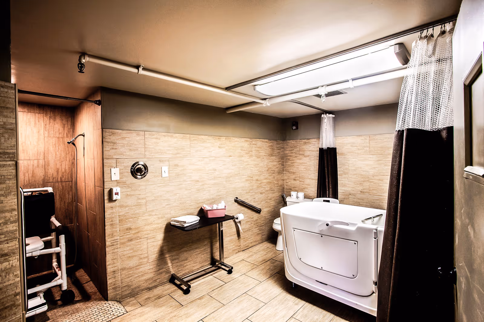 A spacious, accessible bathroom with beige tiled walls and floor. The room features a walk-in bathtub with a safety handle, a toilet, a small table with towels and a tissue box, and a shower area with a shower chair. There are two privacy curtains hanging from the ceiling and a ceiling light providing illumination.